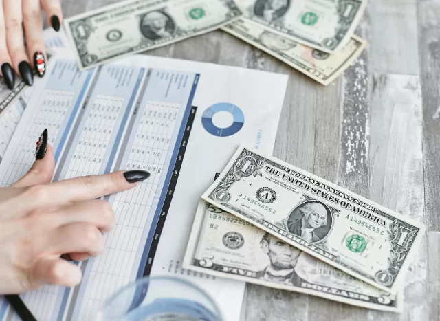 A hand with manicured nails points to a financial table while reviewing Project Budget Management data with US currency nearby.