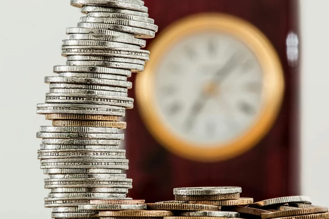 A tall, vertical stack of South African coins next to a blurred clock, illustrating the concept of Project Cost Management.