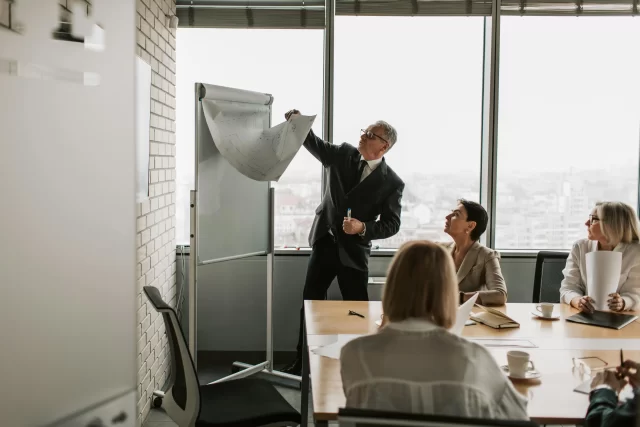 A male executive presenting strategy to his team while Consulting on Management in a high-rise office.