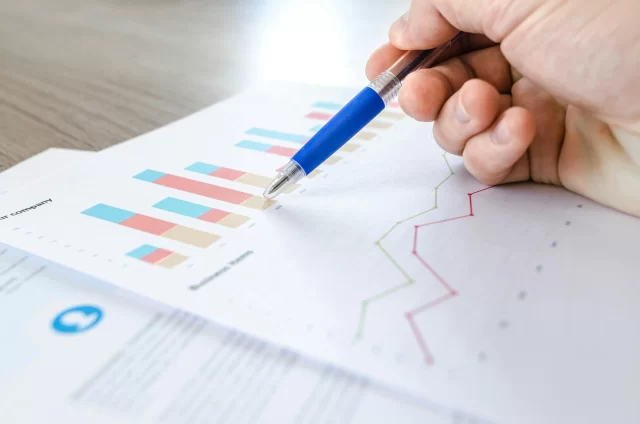 A hand holds a pen pointing to a graph on a NORSTRAT business performance report.