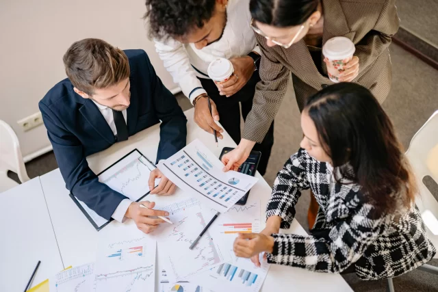 A diverse team of professionals and an implementation consultant reviewing financial reports and data charts in a boardroom.